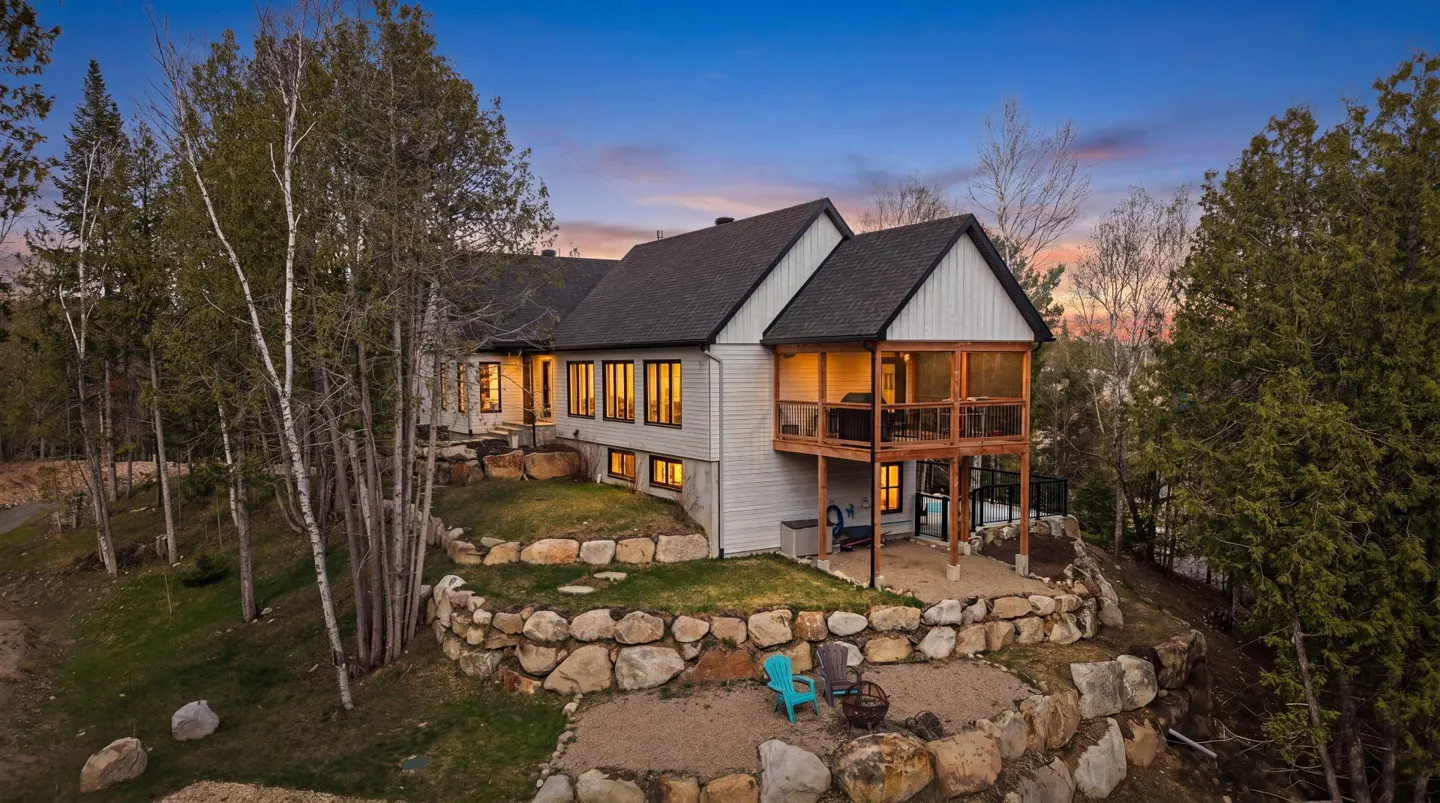 Exterior view of a modern farmhouse with a dark roof, white siding, and a screened-in porch, surrounded by trees and a stone retaining wall.