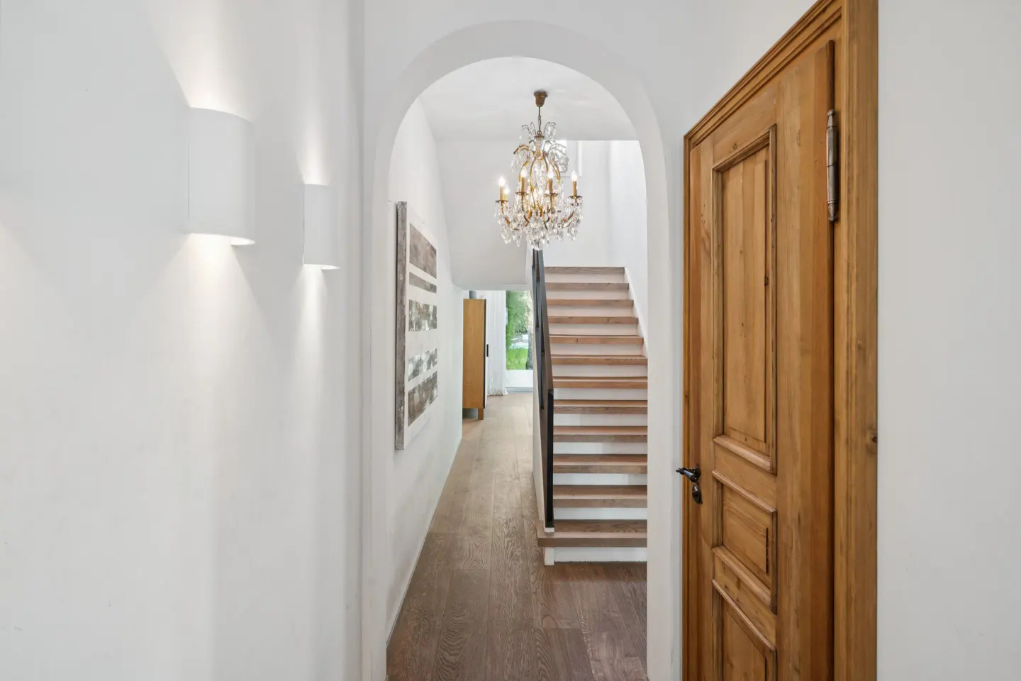Hallway with white walls, wood floors, and a staircase visible through an arched opening. A chandelier hangs above the stairs. A wooden door is on the right.