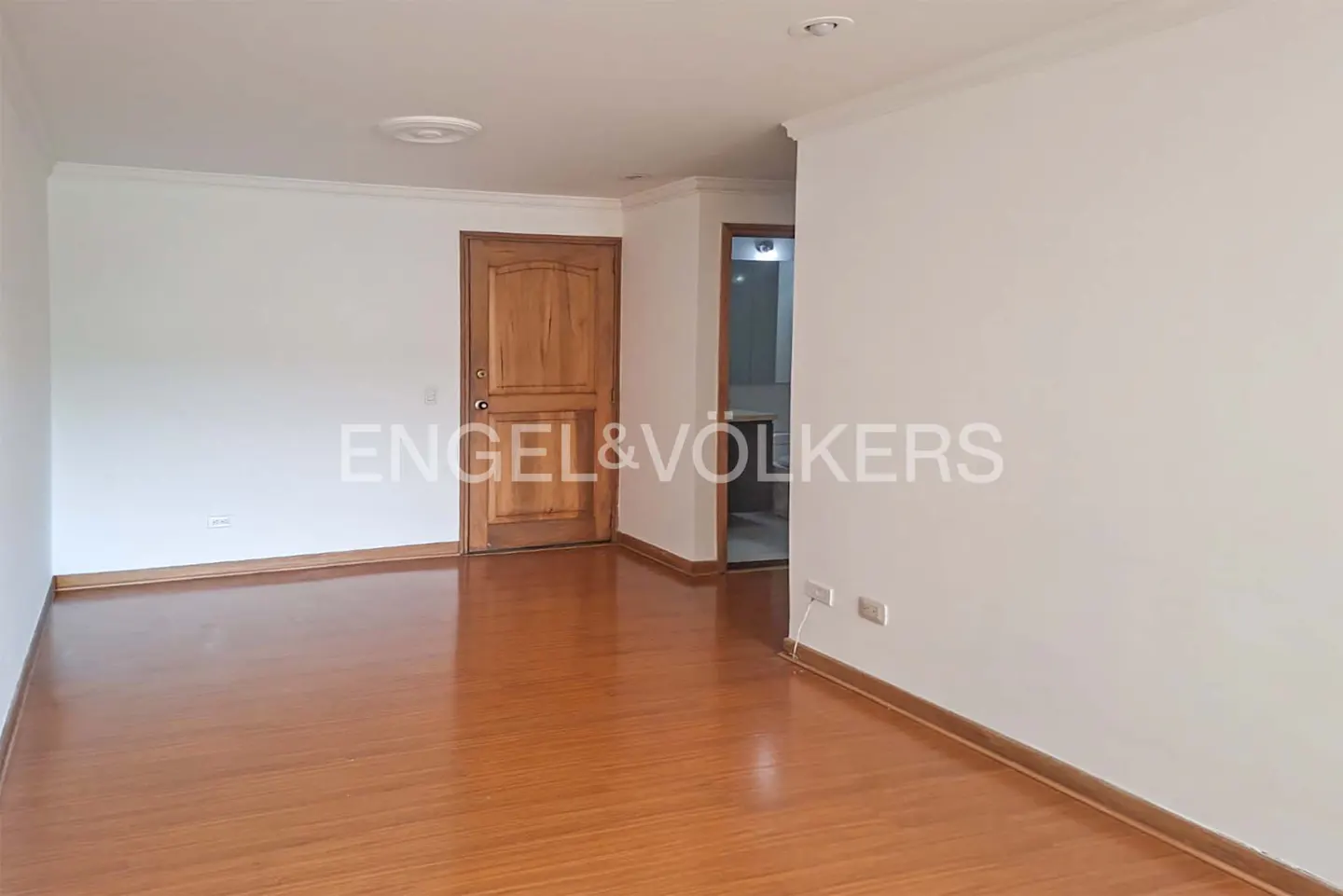 An empty room with white walls, a wooden door, and a shiny wooden floor. A bathroom is visible through a doorway.
