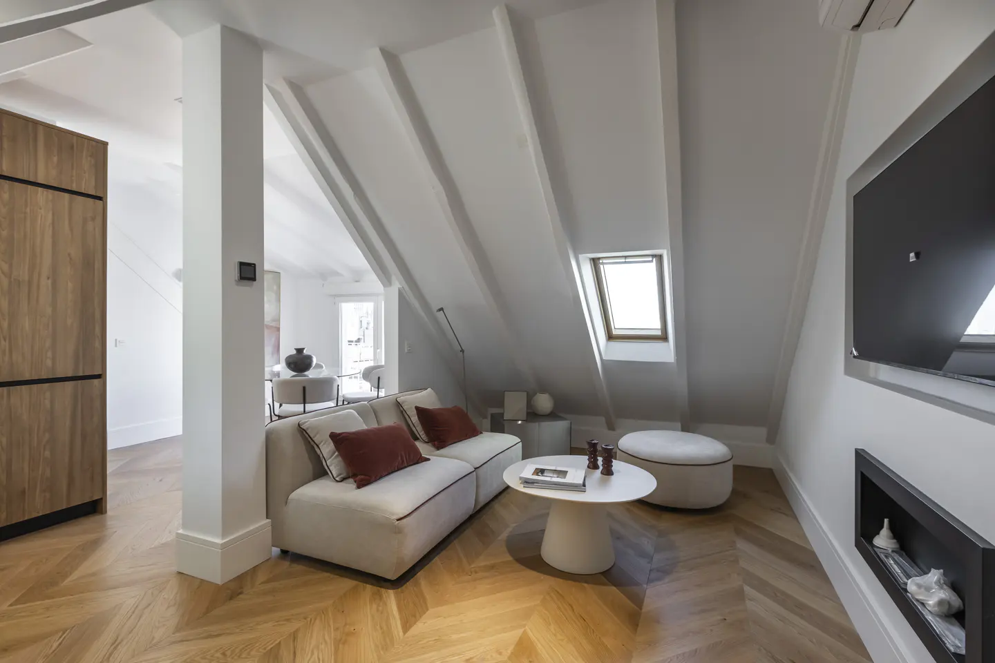 Bright living room with white walls, wood floors, and a beige sofa with red pillows. A skylight is visible in the angled ceiling.