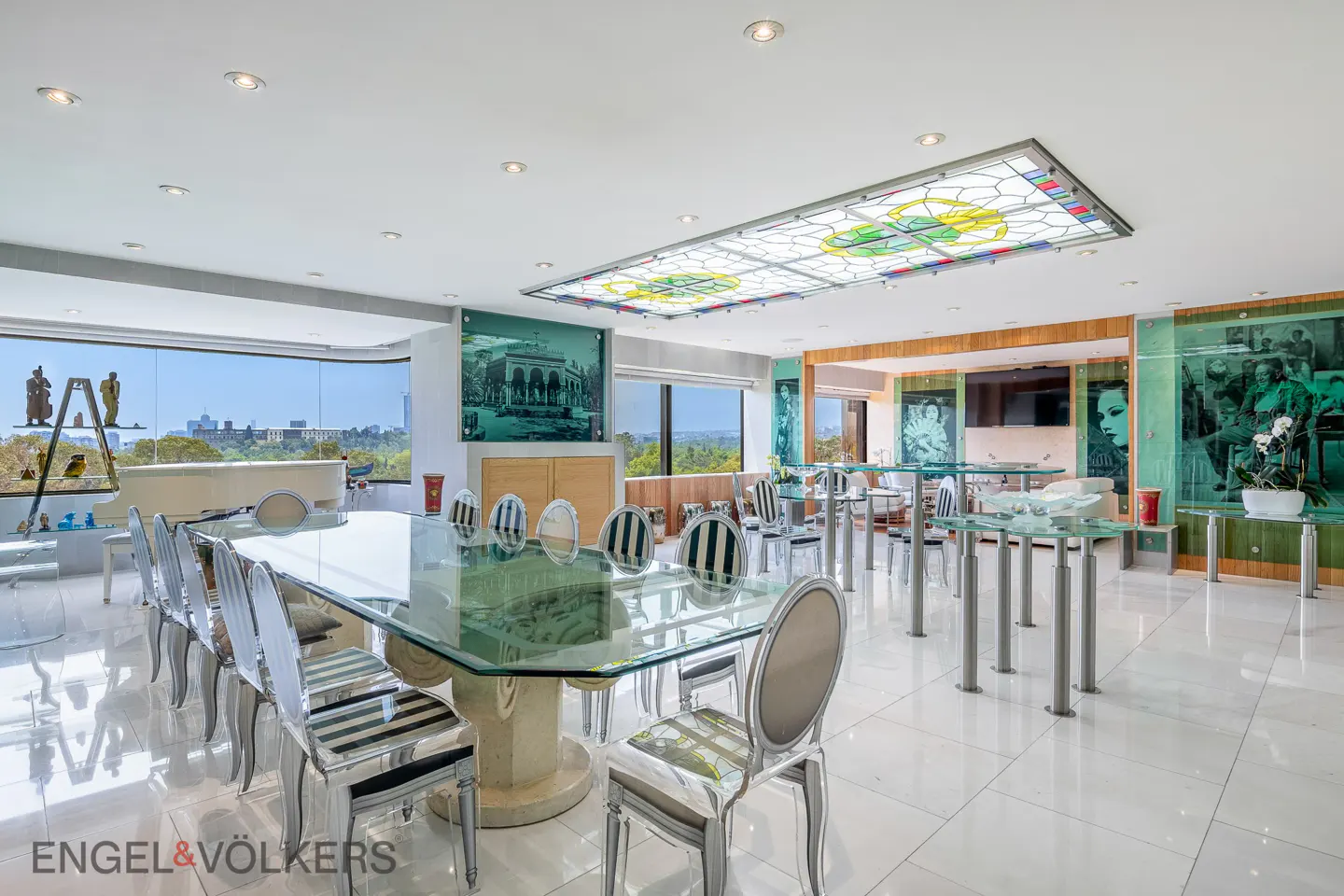 Bright, modern dining room with a glass table, striped chairs, and a stained glass ceiling panel. Large windows offer a city view.