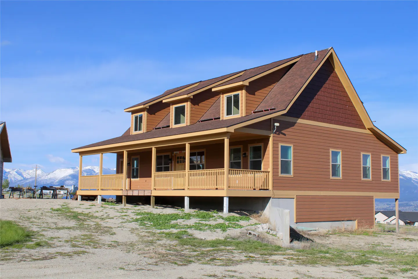Two-story brown house with a porch and a brown roof against a blue sky. Snow-capped mountains are visible in the background.