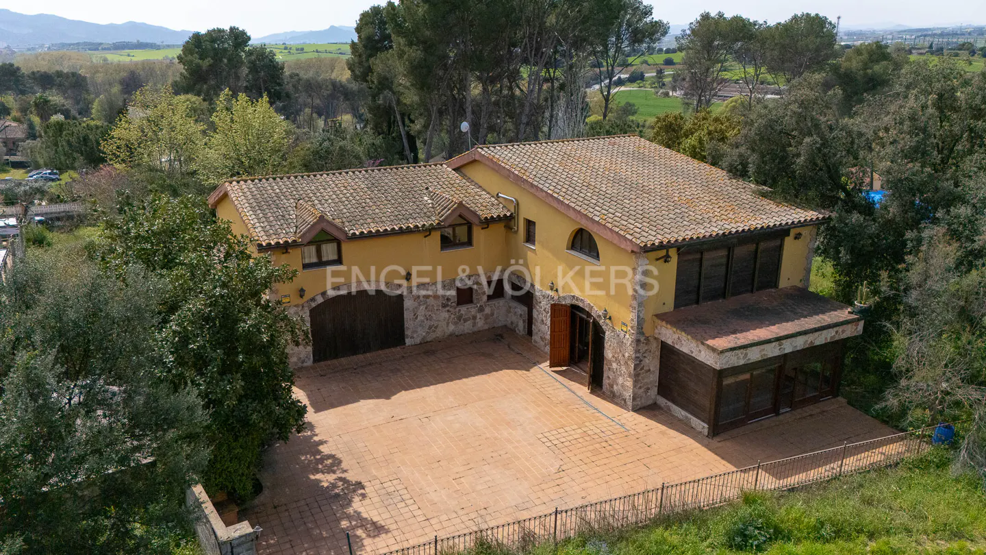 Aerial view of a yellow, stone-accented house with a terracotta tile roof and large brick patio, surrounded by green trees.