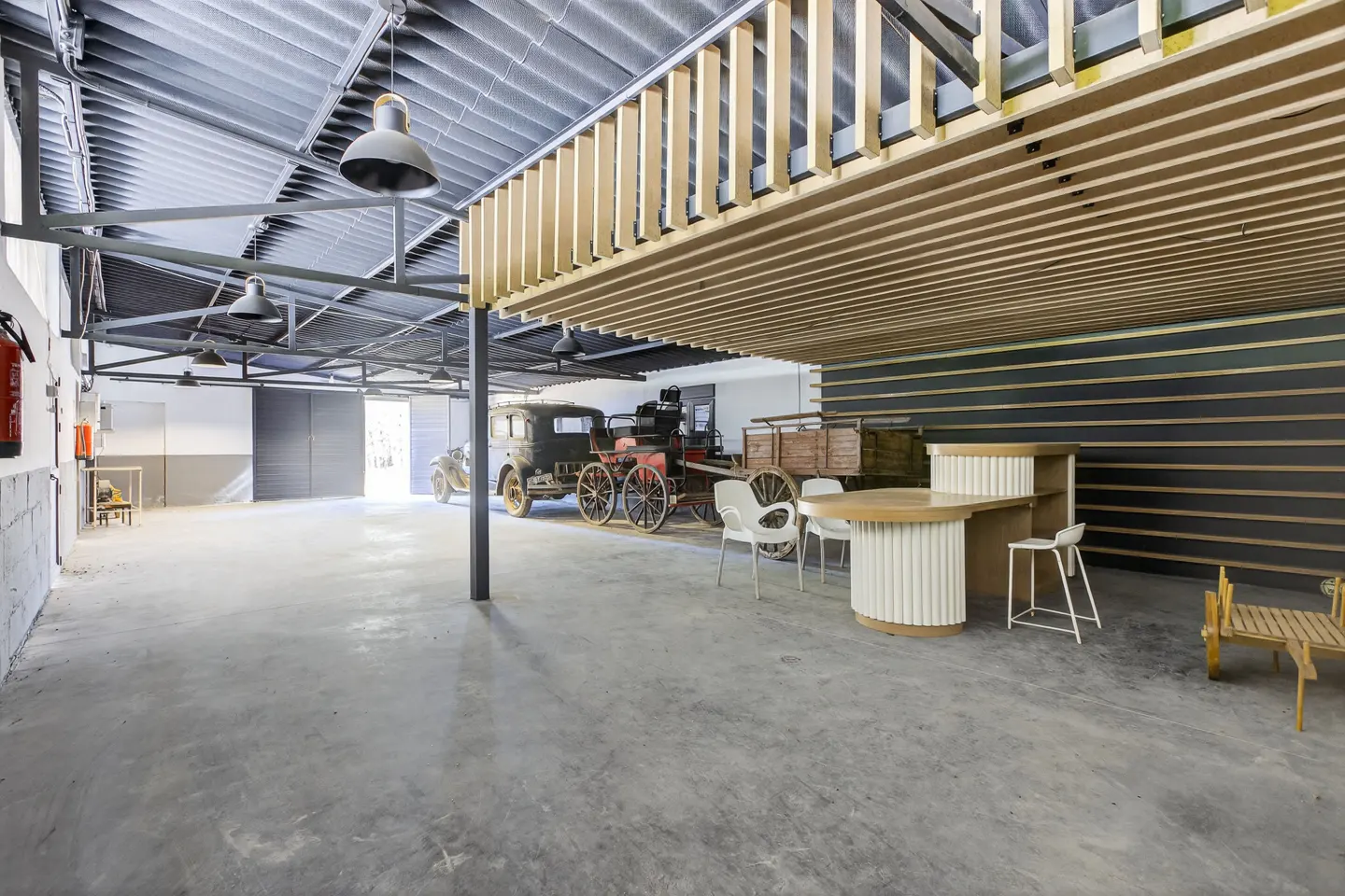 Wide shot of a garage-style space with vintage cars, a horse-drawn carriage, and a modern bar area with white chairs.