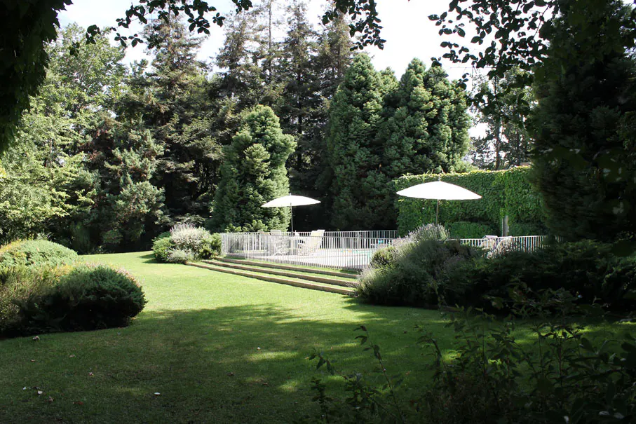 Lush green lawn with trees, bushes, and a pool area with white umbrellas and fence.
