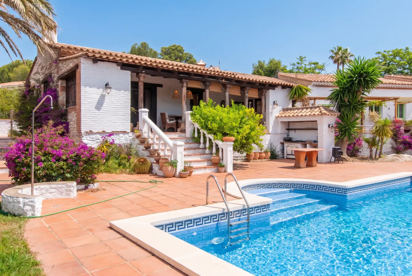 Exterior view of a white brick house with a pool, terracotta patio, and lush greenery under a clear blue sky.