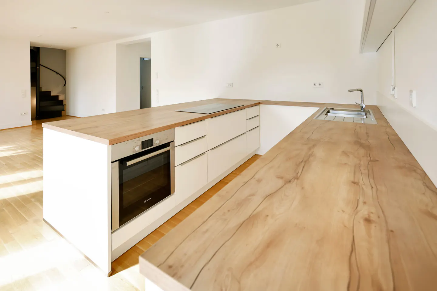 Bright, modern kitchen with white cabinets, wood countertops, stainless steel sink, and oven. A black spiral staircase is visible in the background.