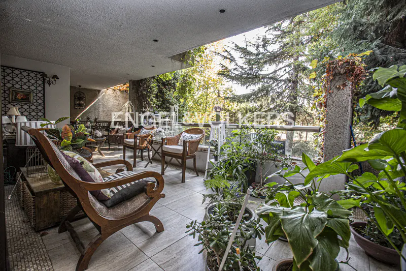 A covered patio with wicker chairs, potted plants, and a view of lush greenery. The floor is tiled, and the space is well-lit.
