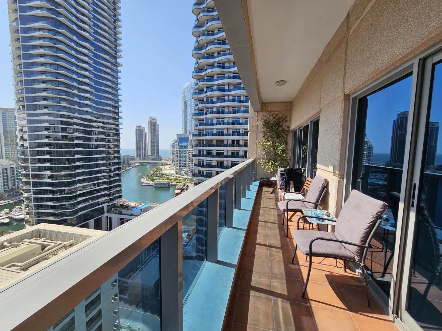 Balcony view of Dubai Marina with chairs, table, and skyscrapers under a blue sky.