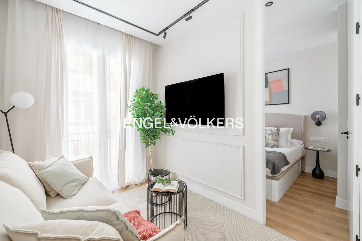 Living room with a cream sofa, TV, and a view into the bedroom with a bed and nightstand.