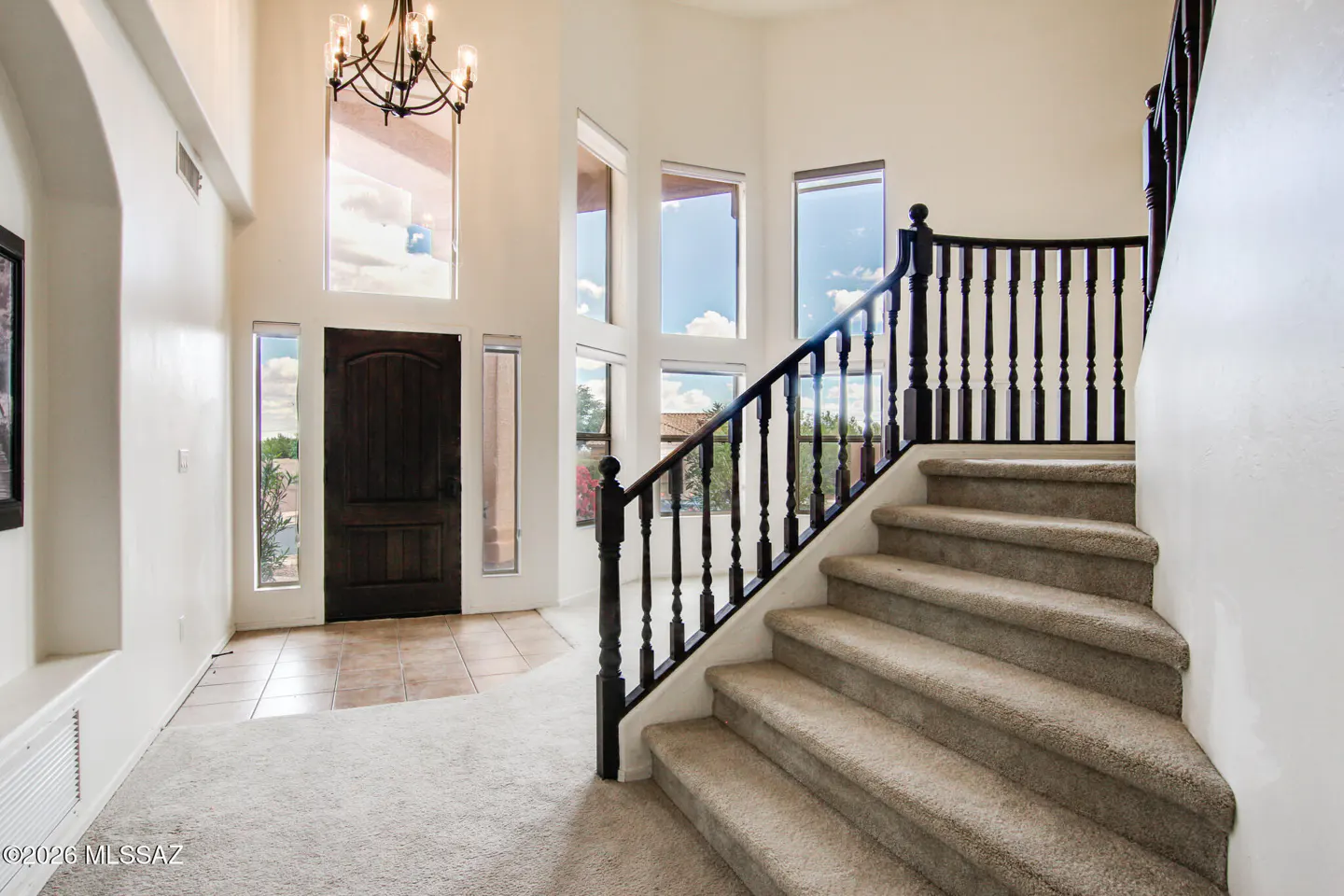 Bright foyer with a dark wood door, tall windows, and a carpeted staircase with a dark wood railing. A chandelier hangs from the high ceiling.