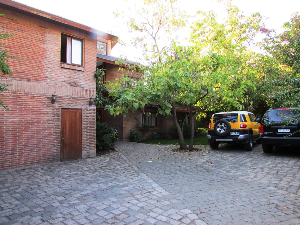Brick house with a wooden door, cobblestone driveway, and two parked cars, one yellow and one black, surrounded by green trees.