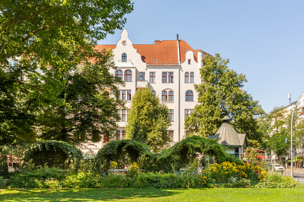 Apartment building with red roof tiles behind a green park with trees and flowers on a sunny day.