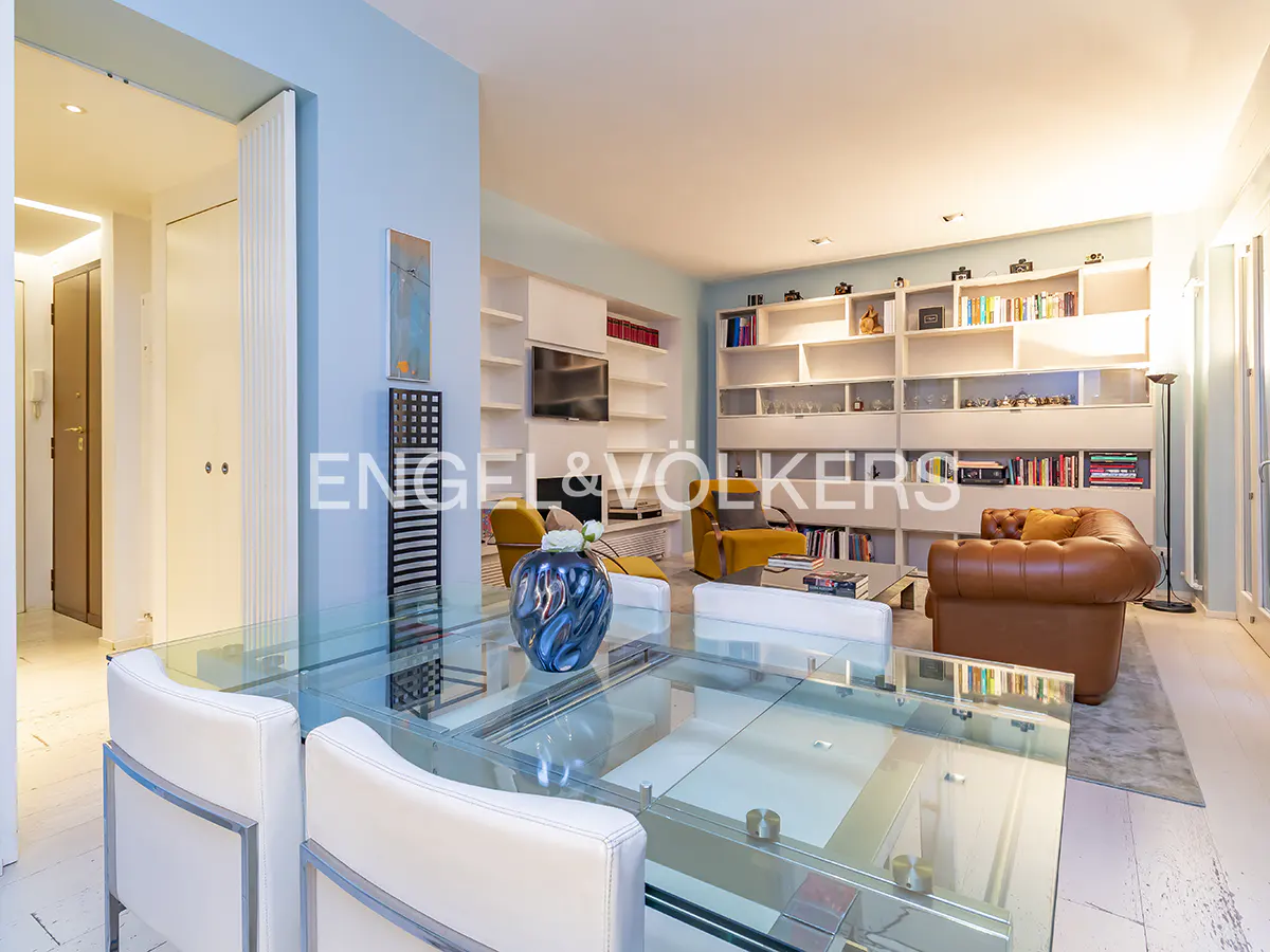 Bright living room with a glass table, white chairs, and a blue vase. Bookshelves line the back wall, with a brown leather couch and yellow chairs.