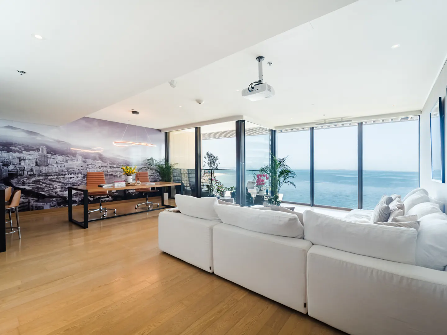 Bright, modern living room with hardwood floors, white sectional sofa, desk with city mural, and ocean view through large windows.