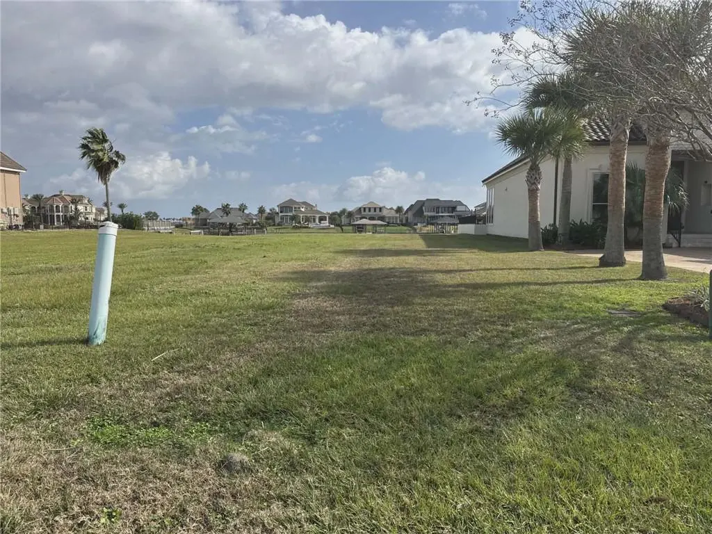 Vacant lot with green grass, a blue pipe, and palm trees. Houses are visible in the background under a cloudy sky.