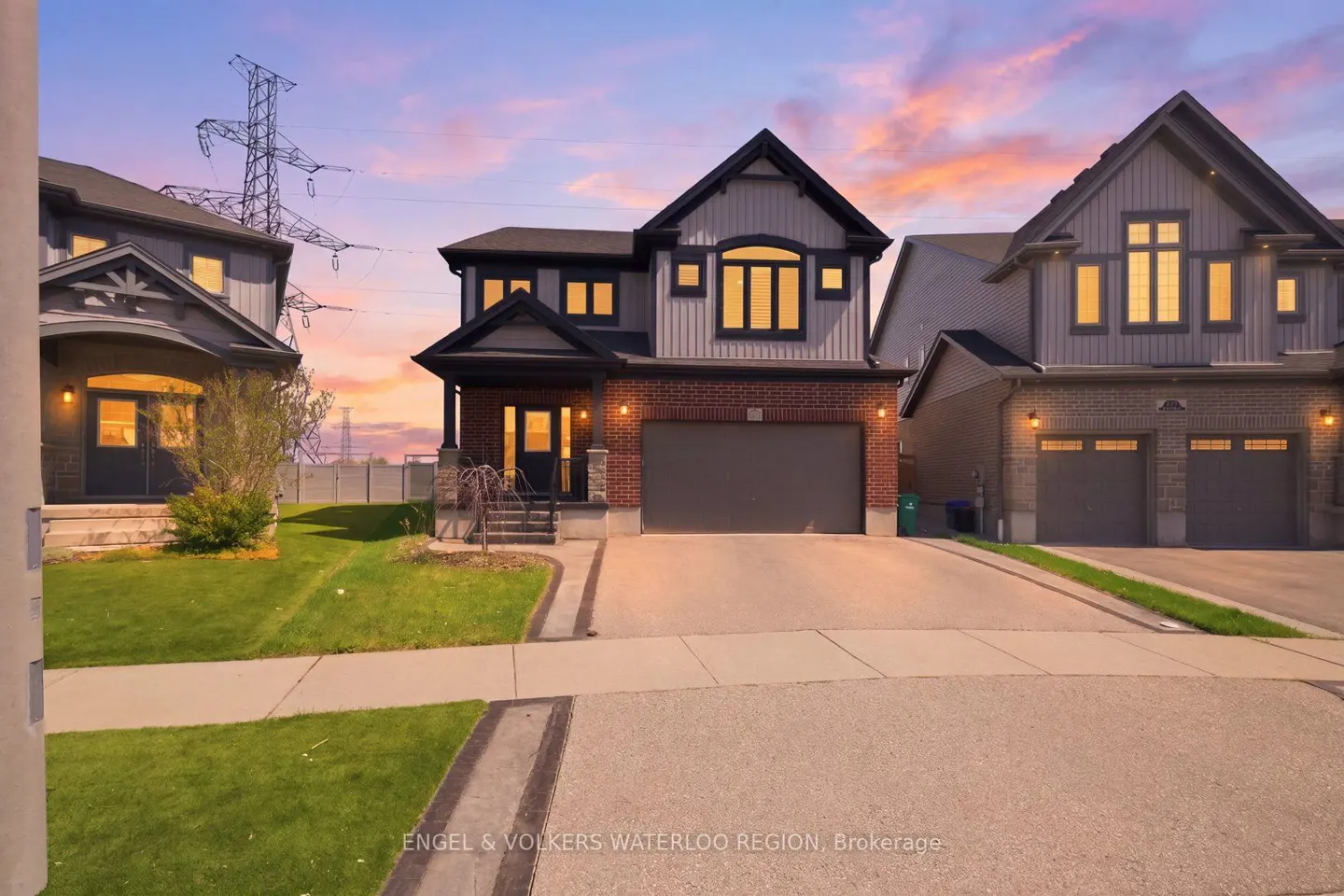 Two-story house with gray siding, brick base, and a gray garage door at dusk. Windows glow with warm light.