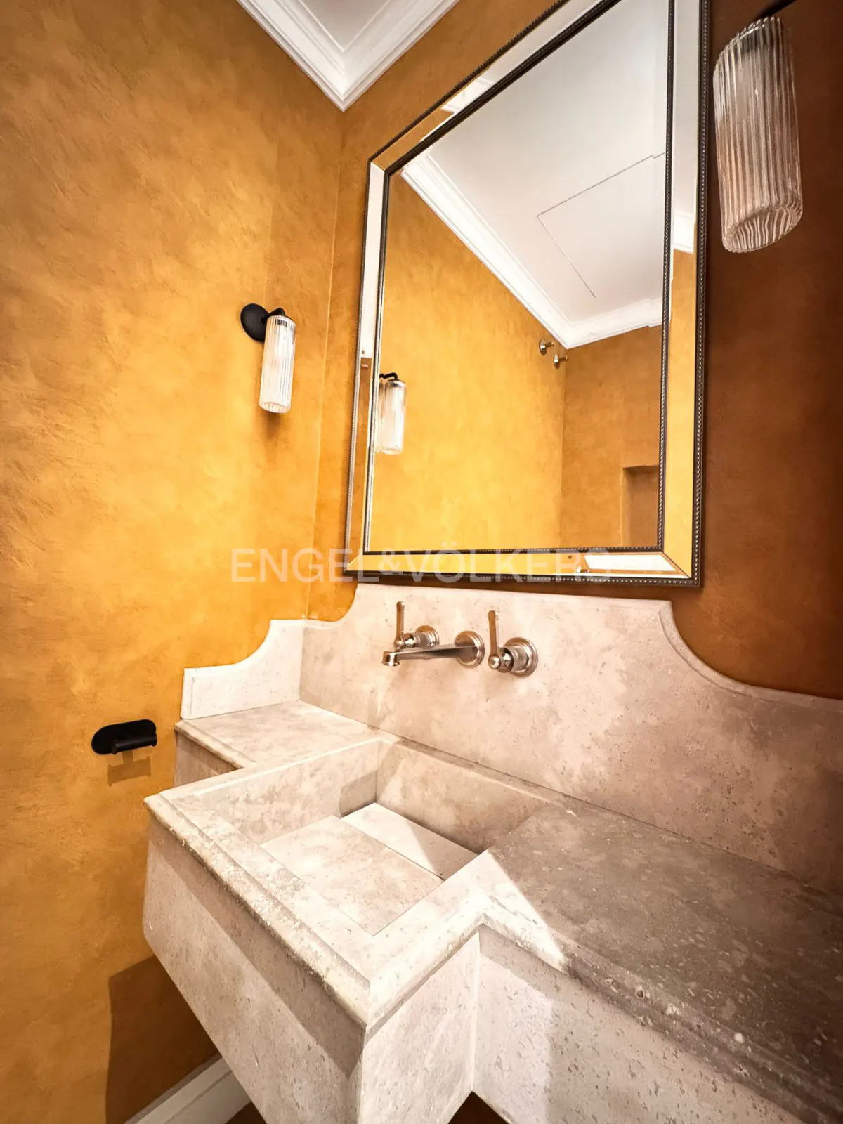 Bathroom with gold walls, a stone sink, and a mirror reflecting the room. A light fixture is on the wall.