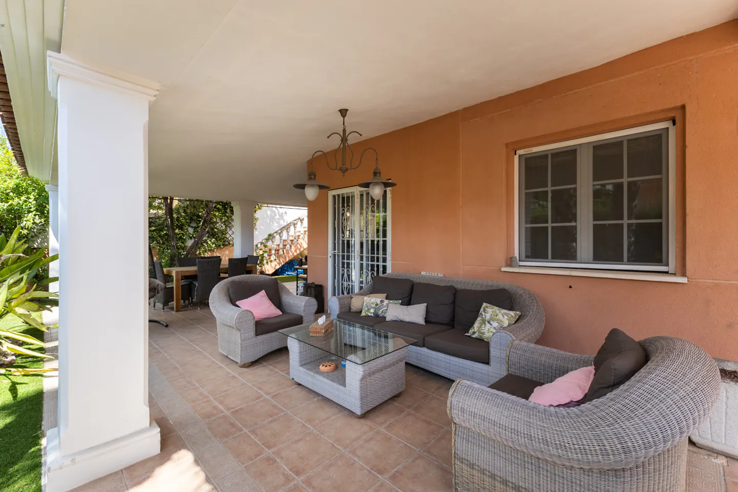 Covered patio with wicker furniture set, including sofa, chairs, and glass table. Orange wall with window and tile floor.