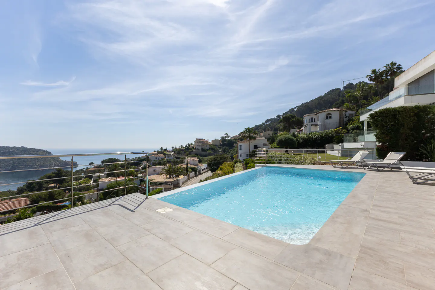 Outdoor pool with blue water, gray tile deck, and metal railing overlooking ocean and hillside homes under a blue sky.