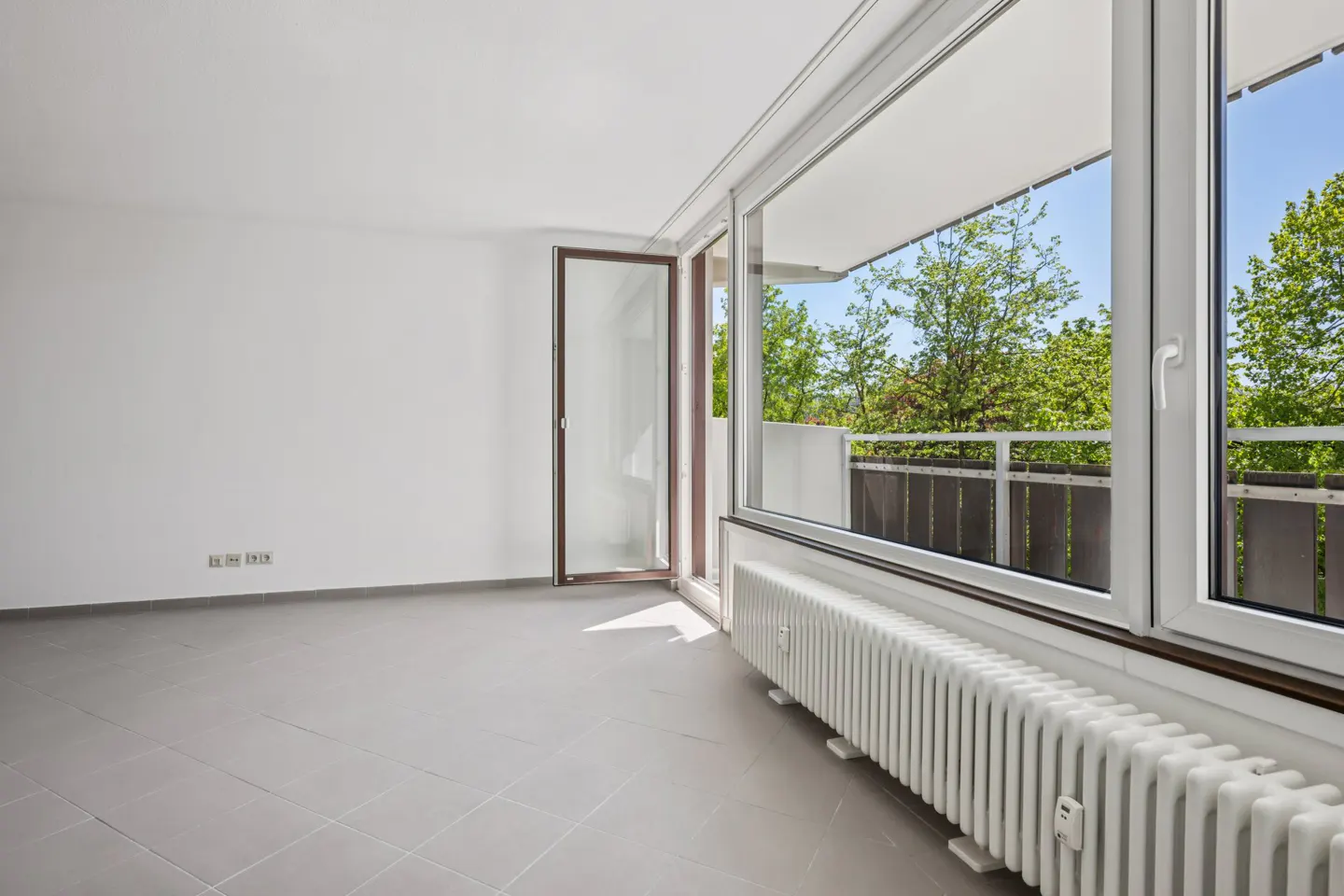 Bright, empty room with gray tile floor, white walls, and radiator. Open glass doors lead to a balcony with green trees visible.