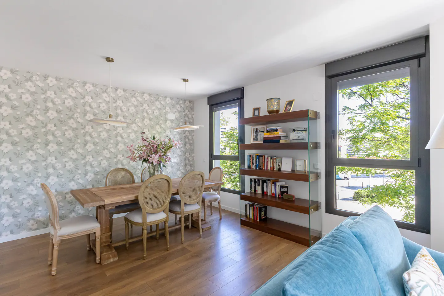 Bright dining room with a wooden table, cane chairs, and floral wallpaper. A bookshelf stands near a window with a view of green trees.