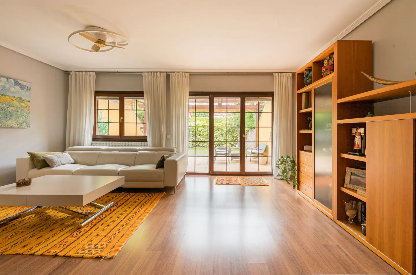 Living room with a white sofa, a light wood bookshelf, and a large window overlooking a green garden.