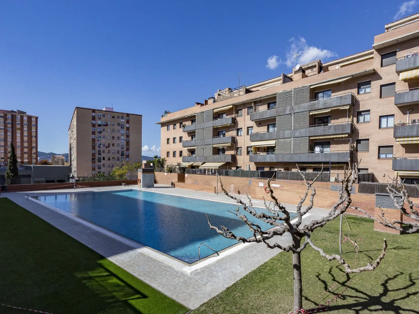 A rectangular swimming pool sits in a courtyard surrounded by a grassy lawn and multi-story brick apartment buildings.