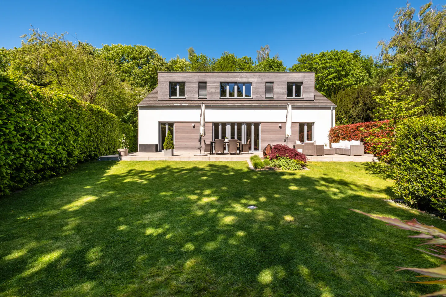 Two-story house with a green lawn, patio furniture, and tall green hedges under a blue sky.