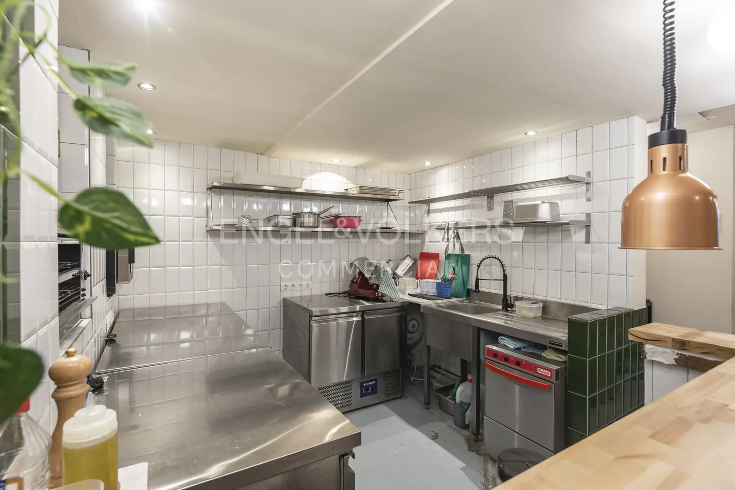 A commercial kitchen with stainless steel counters, white tiled walls, and a copper pendant light.