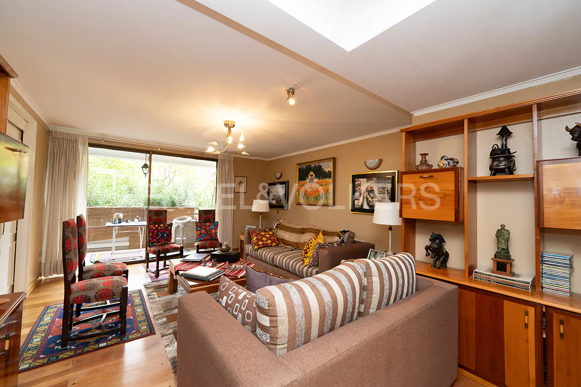 A living room with brown sofas, patterned chairs, and a wooden bookshelf. A balcony is visible through a sliding glass door.