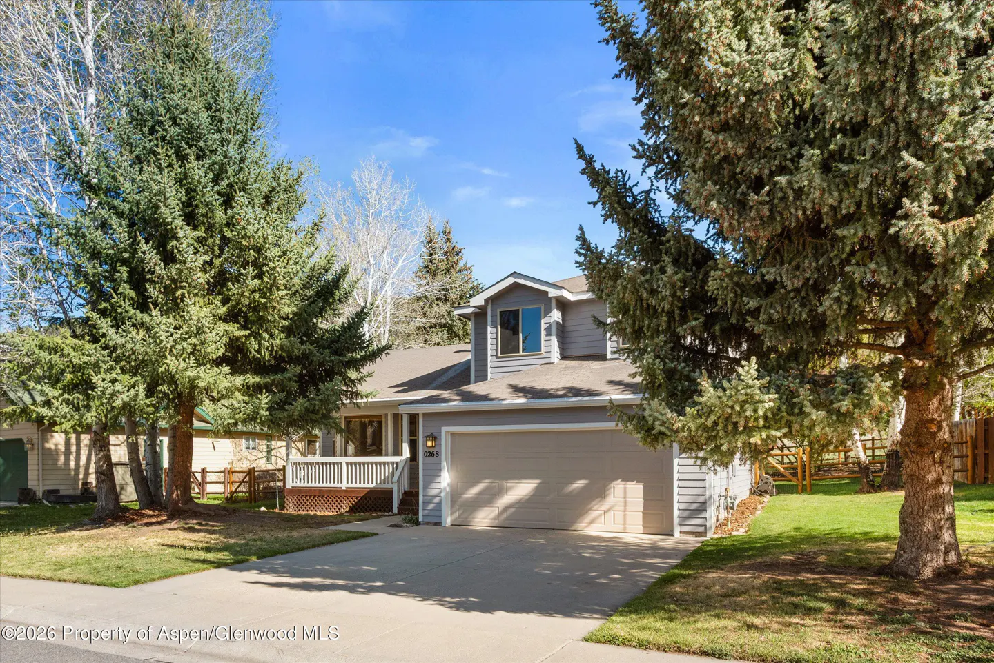 Two-story gray house with a two-car garage, a front porch, and tall green trees on a sunny day.