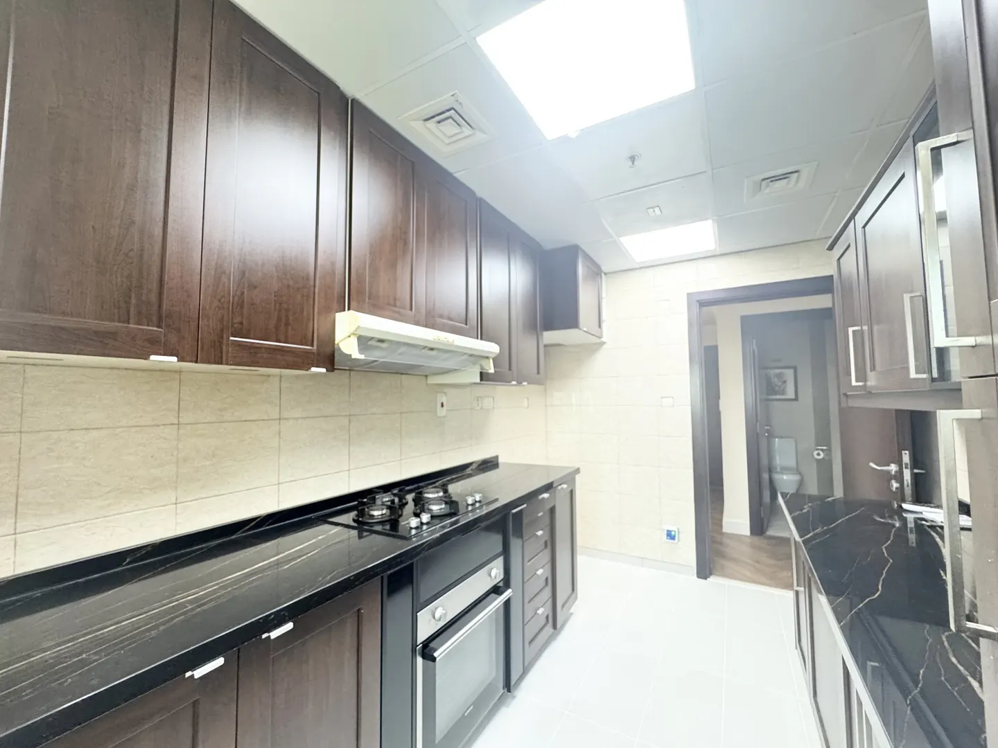 A kitchen with dark wood cabinets, black marble countertops, and stainless steel appliances. A bathroom is visible through a doorway.