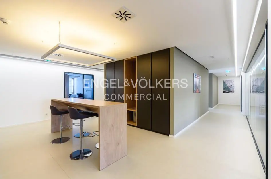 Office space with a light wood table and three black stools. A black cabinet is against the wall. The Engel & Völkers logo is visible.