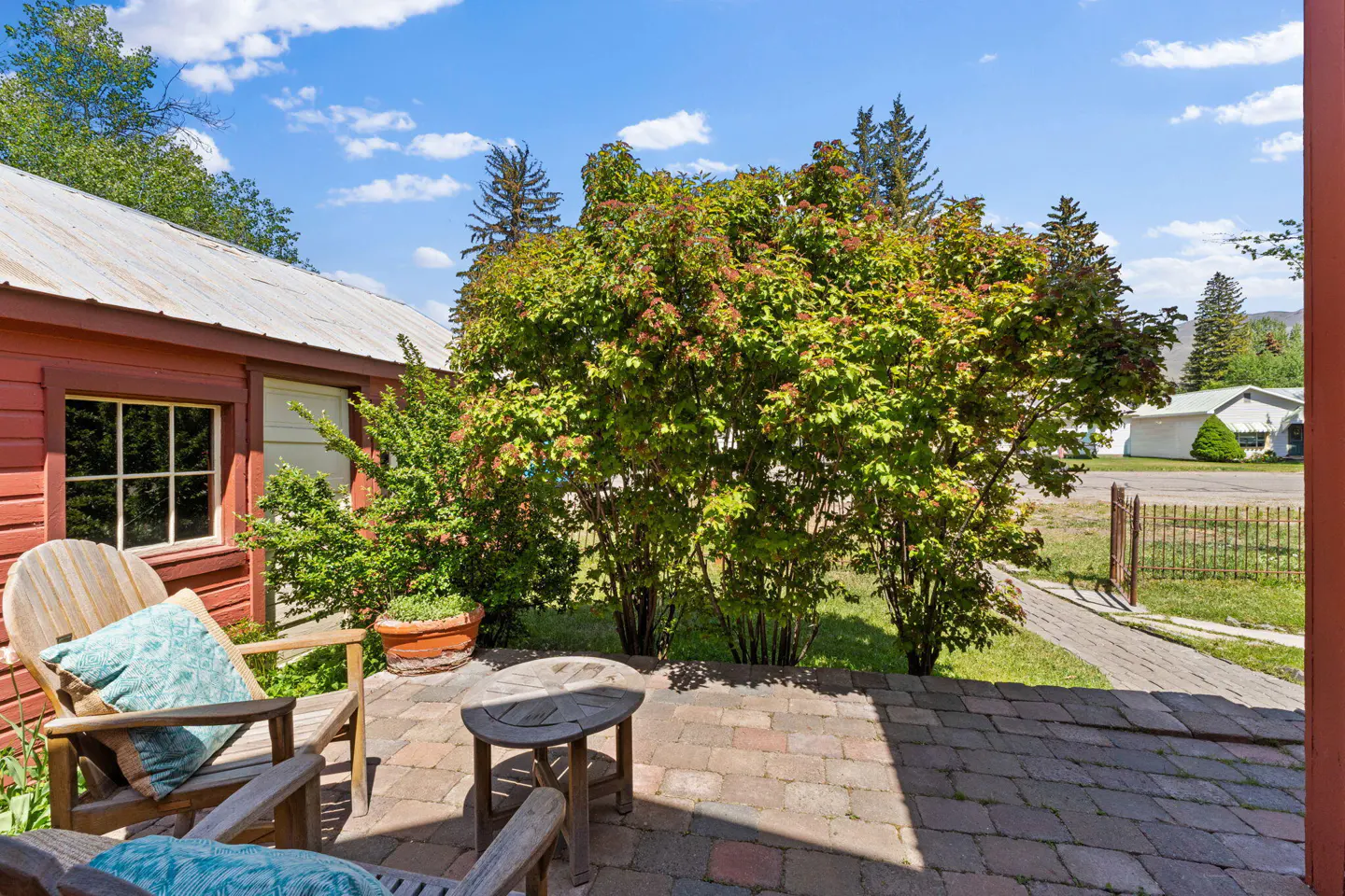 Outdoor patio with brick pavers, wooden chairs with blue pillows, and a small table. A red building and green bushes are in the background.