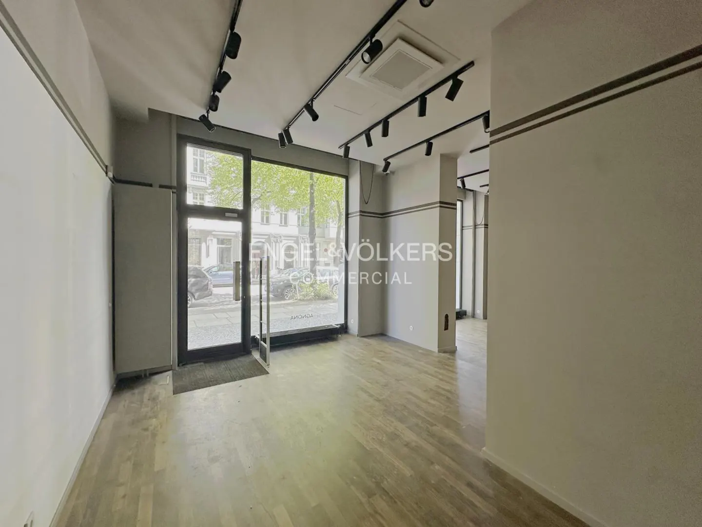 Empty commercial space with wood floors, gray walls, and black track lighting. Engel & Völkers logo on the glass door.
