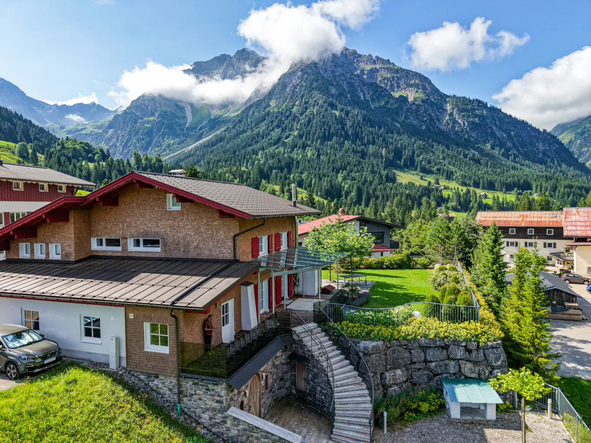 Exterior view of a brown house with red trim, stone stairs, and mountain backdrop.