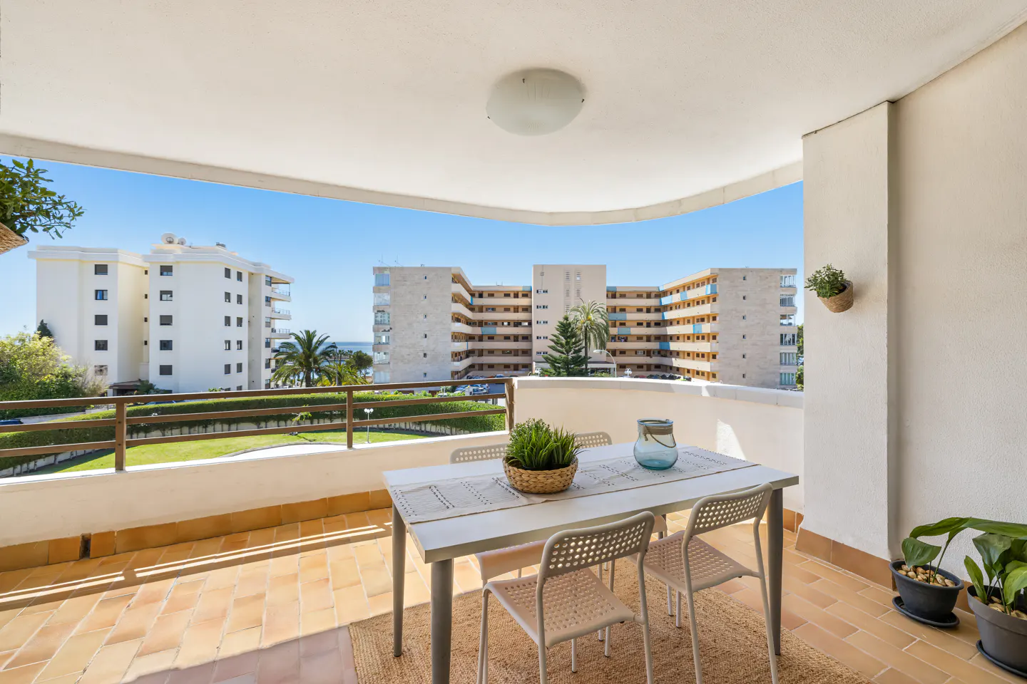 Balcony view with a white table, four chairs, and a plant. Buildings and blue sky are visible in the background.