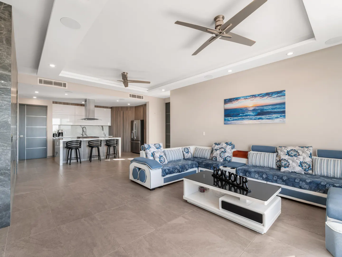 Open-concept living space with a blue and white sectional sofa, kitchen island with stools, and a ceiling fan.