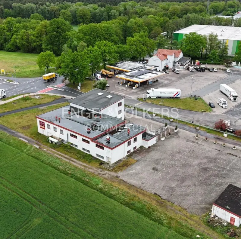 Aerial view of a white commercial building with a gas station and trucks in the background. Green fields and trees surround the property.