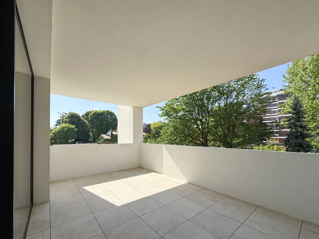 Spacious, empty white balcony with tiled flooring, overlooking lush green trees and a distant apartment building.