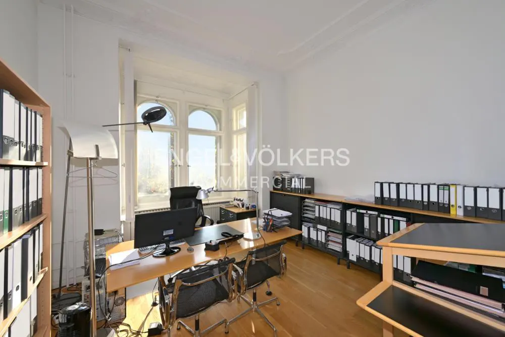 Bright office space with wood floors, a large desk, black chairs, and shelves filled with binders. A tall window lets in natural light.