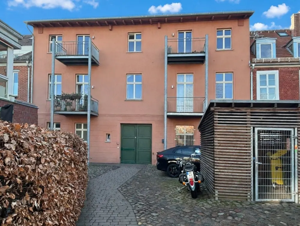 A three-story peach building with balconies, green doors, and a brick walkway. A car and motorcycle are parked nearby.