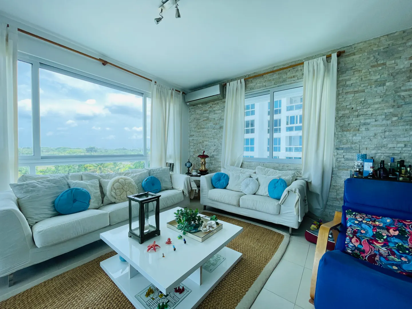 Bright living room with white sofas, blue pillows, and a white coffee table on a jute rug. Large windows offer a view of greenery.