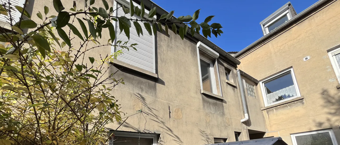 Exterior view of a beige building with white-framed windows, some with closed white shutters, under a blue sky. A leafy bush is in the foreground.