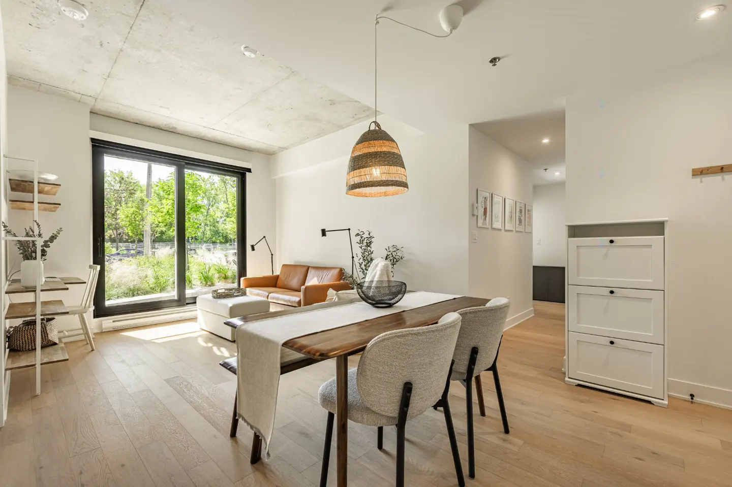 Bright, modern living space with wood floors, a brown leather sofa, and a dining table with gray chairs. A large window overlooks greenery.