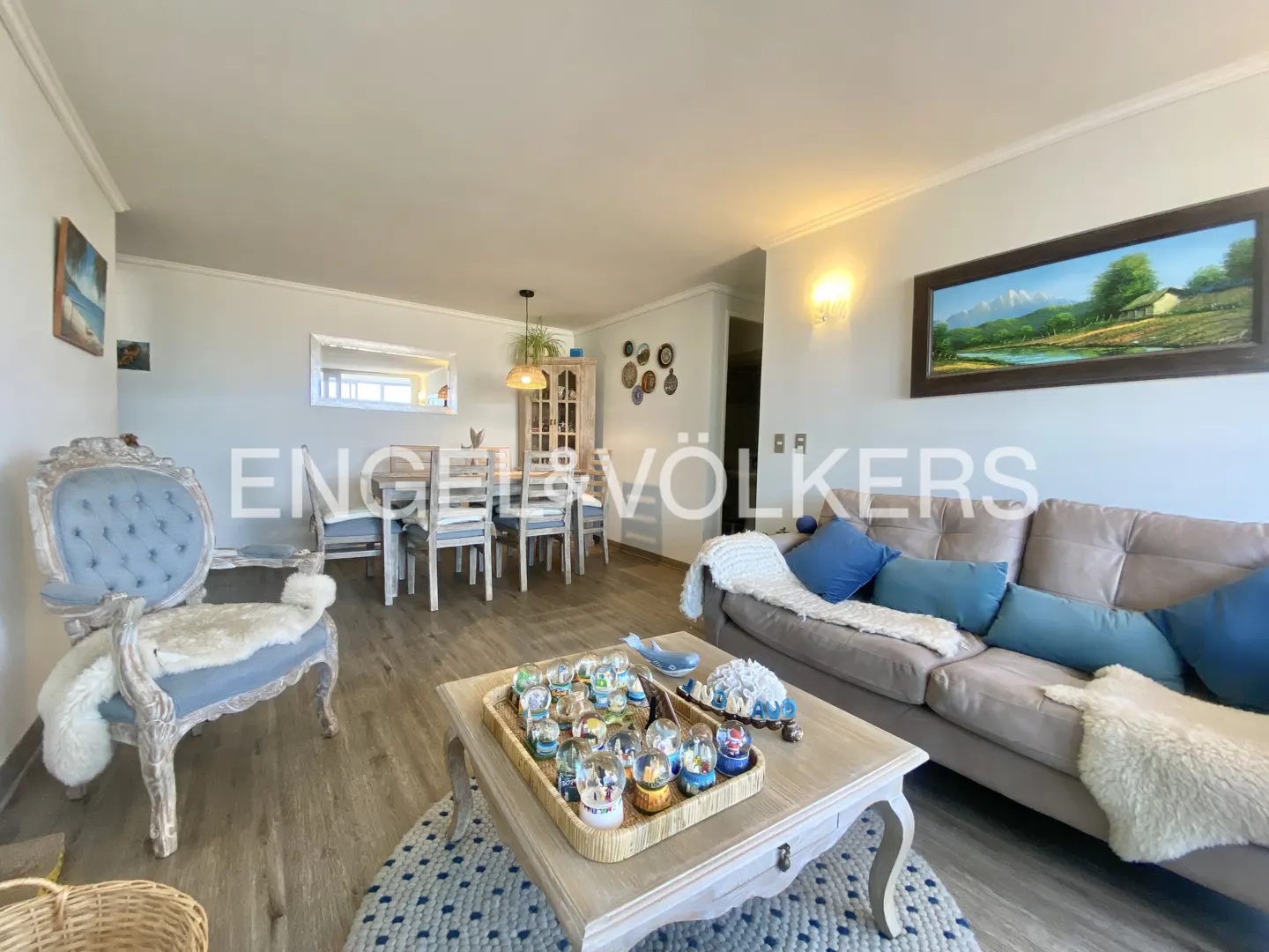 Living room with a gray sofa, blue pillows, a blue chair, and a coffee table with snow globes. A dining table is in the background.