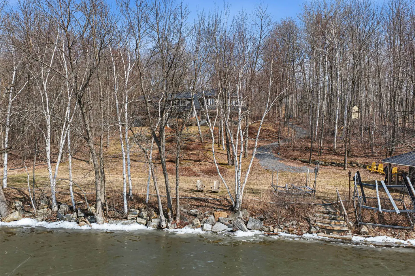 Lakefront home with a dock and a view of the water. The house is surrounded by trees and has a gravel driveway.