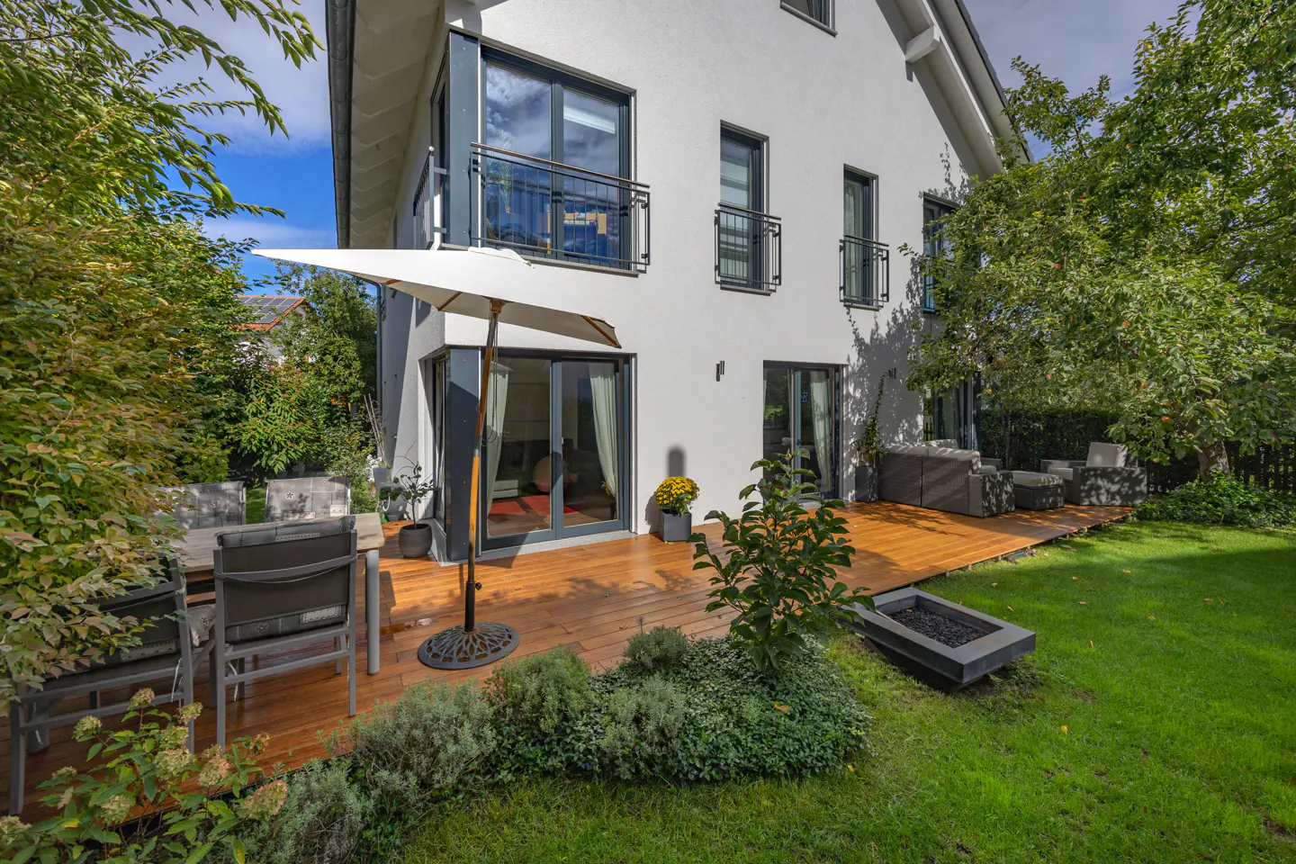 Modern white house with a wooden deck, outdoor dining set, patio furniture, and a green lawn on a sunny day.