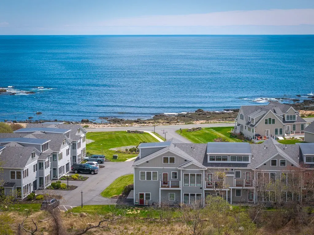 Coastal real estate view: Gray townhouses with ocean backdrop under a blue sky. Cars parked in driveways.